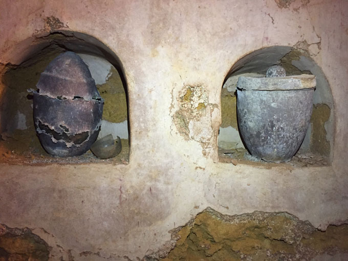 A mauseoleum wall with two alcoves. The left alcove contains an egg-shaped container, and the right alcove contains a bucket-shaped urn.