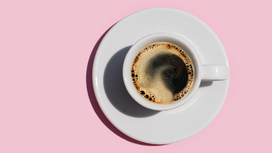 An over head photo of a mug of coffee sitting on a white plate all sitting on a pink background.