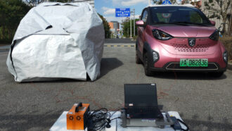 article material science A photo of two cars parked next to each other. The car on the left is covered by a large white tarp that is wrapped around it while the car on the right is pink and exposed.