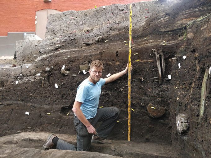 photo of Søren Sindbæk kneeling in an excavated trench and holding a measuring table to show the full height of the trench