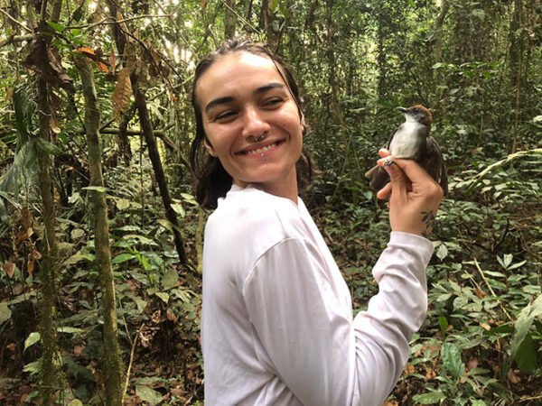 A woman standing in a rainforest, smiling and holding a fire-crested alethe, a small bird with a reddish-brown crest on its head