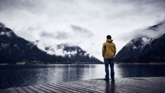 A photo of a man in a yellow raincoat standing at the end of a dock with his back to the camera. The background, in black and white, shows Lake Achen in Austria.