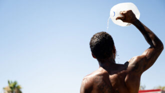 A photo of a man facing away from the camera and pouring water out of a 2-liter jug onto his head.