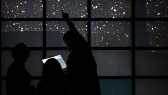A photo of three people, seen in silhouette, pointing at stars on a large wall of several screens.