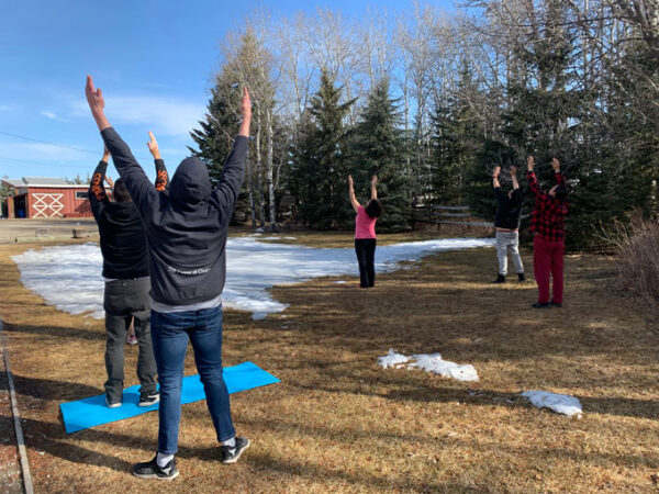 A photo of participants in a land-based healing program in rural Alberta, Canada engaging in a morning prayer meditation. Five people standing with their legs shoulder-width apart and holding their arms up to the sun.