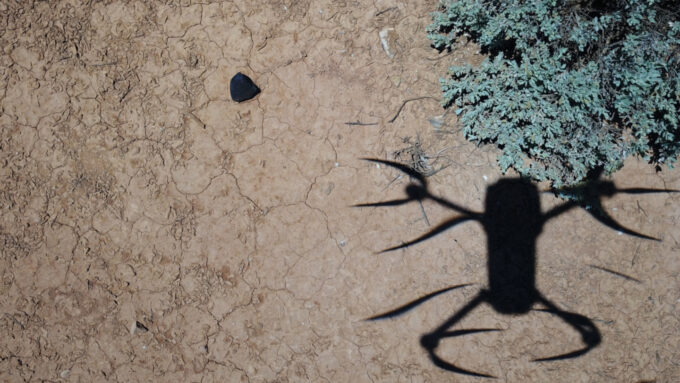 A drone casts a shadow on the ground while it hovers above a dark object flagged as a potential meteorite.