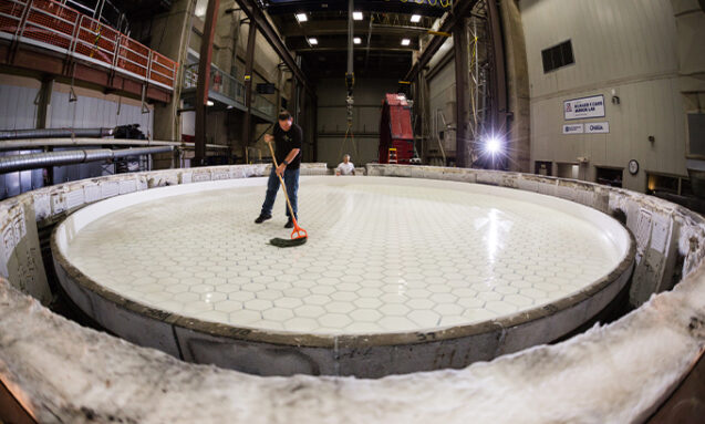 A photo of a man with a mop standing on top of the smooth, clear surface of the glass that will become the Ginat Magellan Telescope's fifth mirror.
