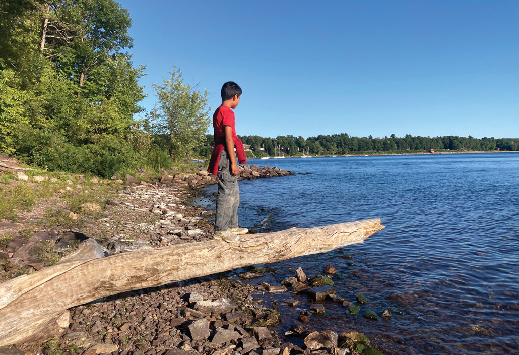 A photo of a young boy standing on a log that is hanging over a body of water. Trees line the body of water's shore.