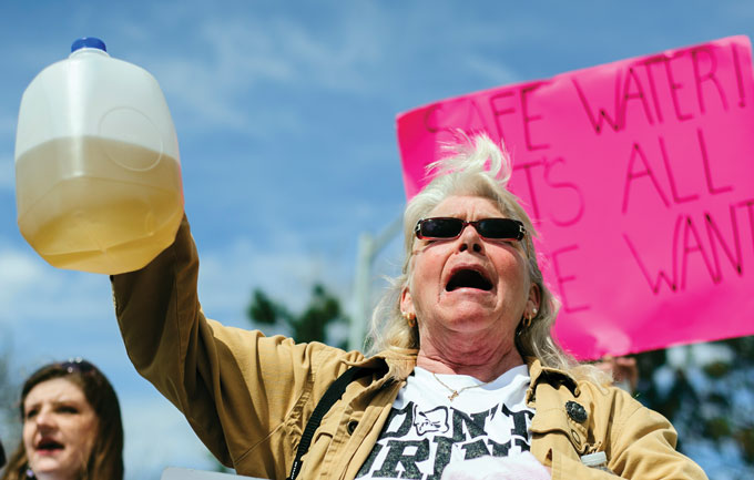 A person holds a jug of brown water with a pink &quot;Safe Water&quot; sign in the background.