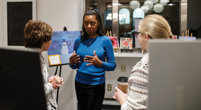 A person stands with two others in a room and paintings, one of a snowman, surround them.