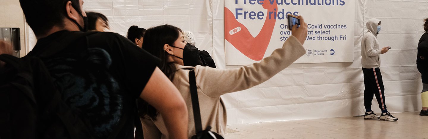 photo of six or so people in Grand Central Station wearing facemasks. a woman in the center takes a selfie
