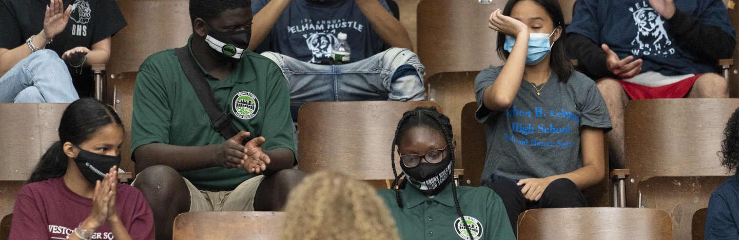 A small group of students in a lecture hall, wearing masks, with empty chairs between them. Several are applauding