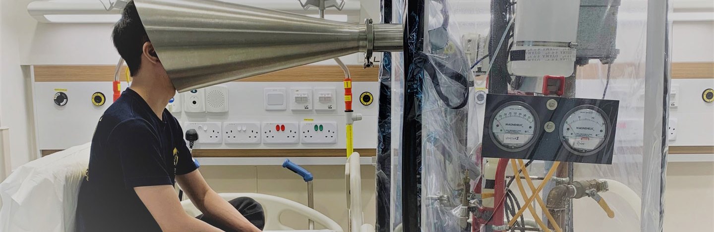 a researcher sits crosslegged on a hospital bed with their face inside a silver cone that's connected to a transparent chamber full of scientific apparatus