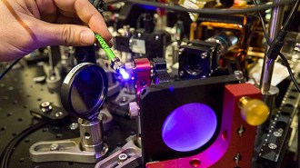 a hand holding a thin green wire at the end of which is a bright light, surrounded by scientific apparatus