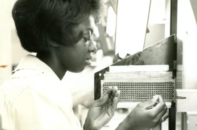 an old, black-and-white photo of a woman threading metal wire through small holes in a machine