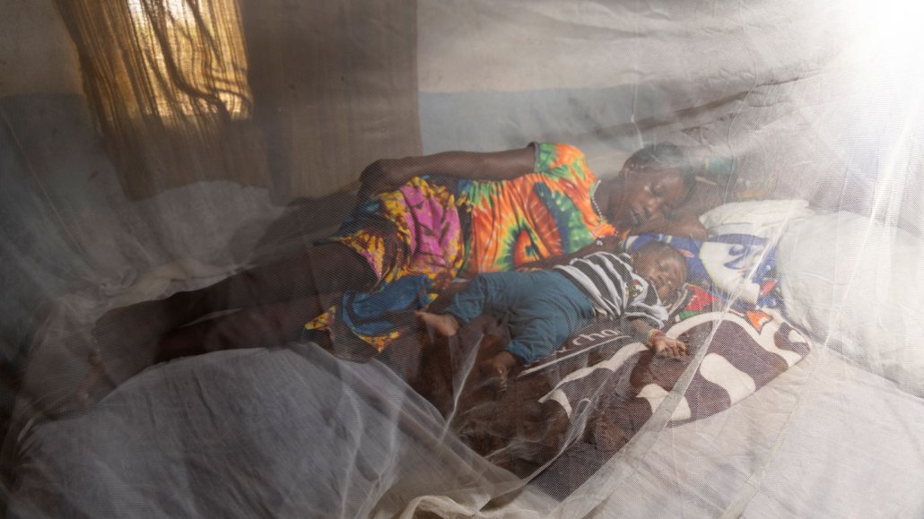 a woman sleeps next to her baby. The photo is taken through a mosquito net.