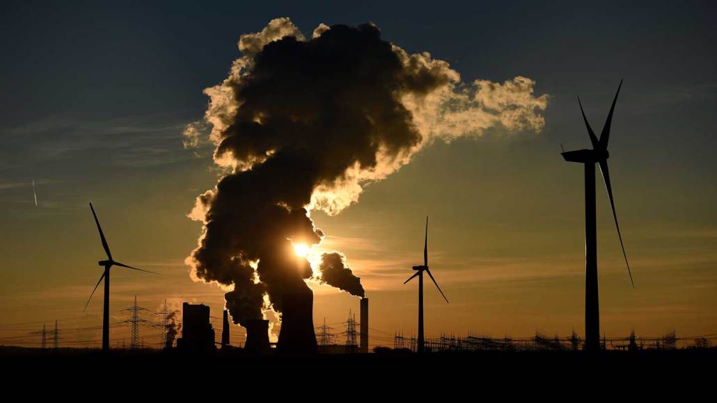 Wind turbines stand near a coal-fired power plant in Germany, with steam rising from the plant's cooling towers.