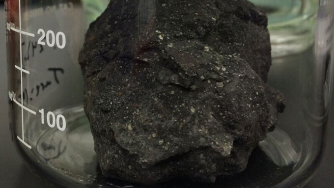 A photo of a two-gram black chunk of meteorite sitting inside a glass beaker