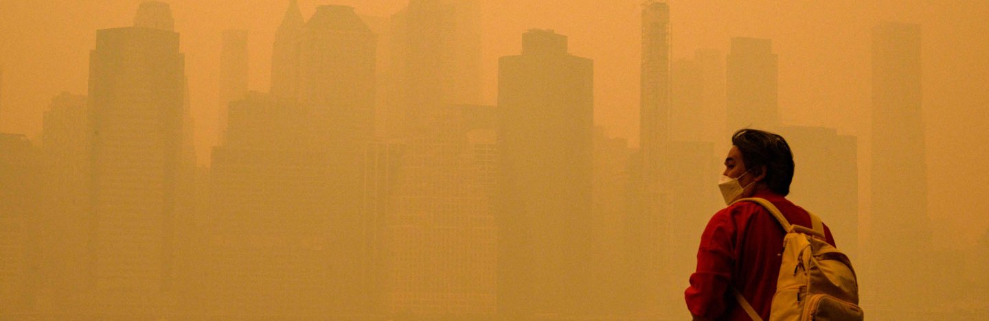 A photo of a person standing in front of the New York City skyline which is barely visible through an orange haze.