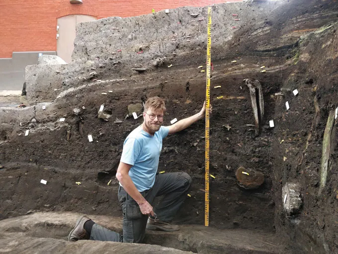 photo of Søren Sindbæk kneeling in an excavated trench and holding a measuring table to show the full height of the trench