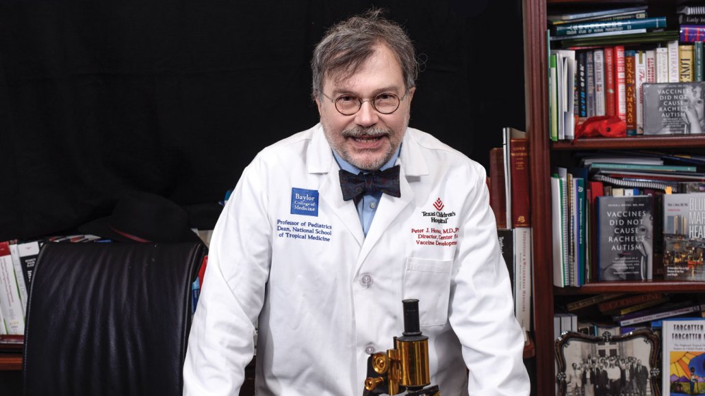 Peter Hotez stands at a desk with a microscope in front of him and a bookshelf in the background.