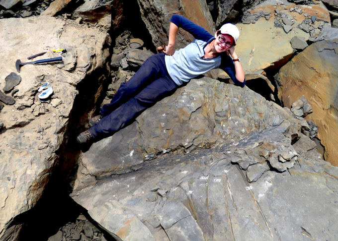 A woman poses horizontally on top of a gray fossil of a tree trunk and extending leaves.