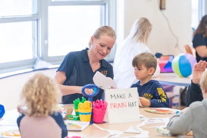 Rain Bosworth interacting with a child sitting next to her. Bosworth's blonde hair is pulled back into a ponytail, and she's wearing a short-sleeve navy shirt that displays the RIT logo in orange. The child. who has short blonde hair and wearing a navy hoodie, is watching Bosworth cutting a piece of paper with a pair of scissors. They are both sitting at a round table, which has a sign that reads, "Make a brain hat."