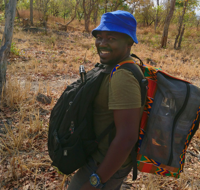 A scientist sports a custom-made backpack that's built to carry wild mosquitoes out of the field. It's cube-shaped, partly mesh, and is edged by colorful fabric. He's carrying a regular backpack on his front.