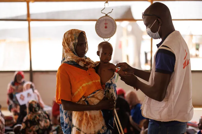A woman in an orange blouse with light scarf wrapped around her head holds a young child. A man wearing a surgical mask wraps a paper tape around the toddler's upper arm to screen for malnutrition.
