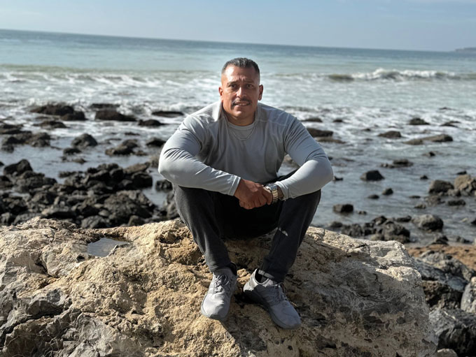 A photo of a man smiling slightly and sitting on a large rock in front of the ocean