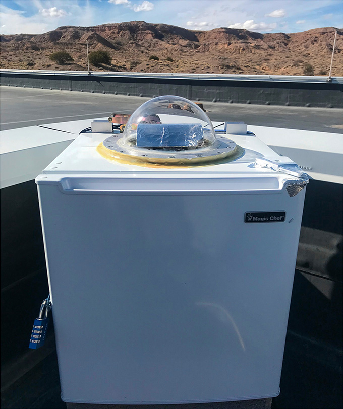 A minifridge with a clear dome on top sits in front of a desert landscape