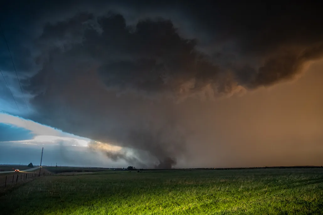 Beneath dark storm clouds, a tornado swirls across farmland.