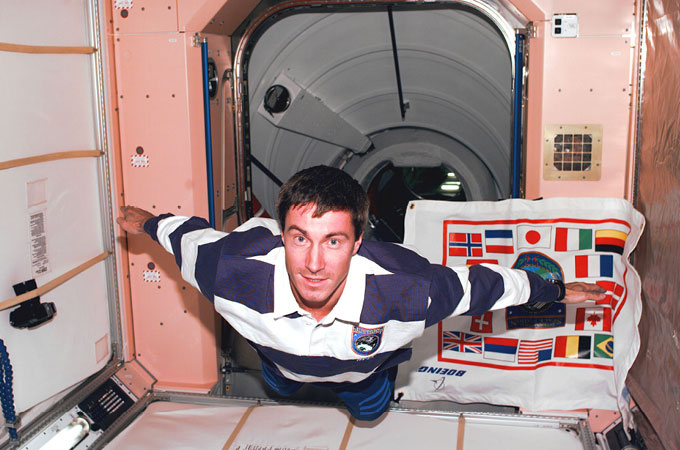 Cosmonaut Sergei Krikalev floats in the International Space Station in front of a poster with many different nations' flags.