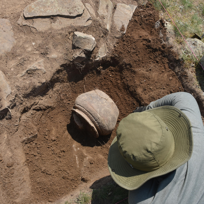A person wearing a green hat crouches over an excavaed hole in which a piece of pottery rests.