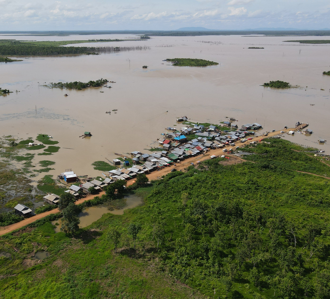 This aerial view of part of the Sesan River, a tributary to the Mekong River, shows a road going out to the river's edge, lined with houses. The river is a muddy expanse dotted with some spots of vegetation.