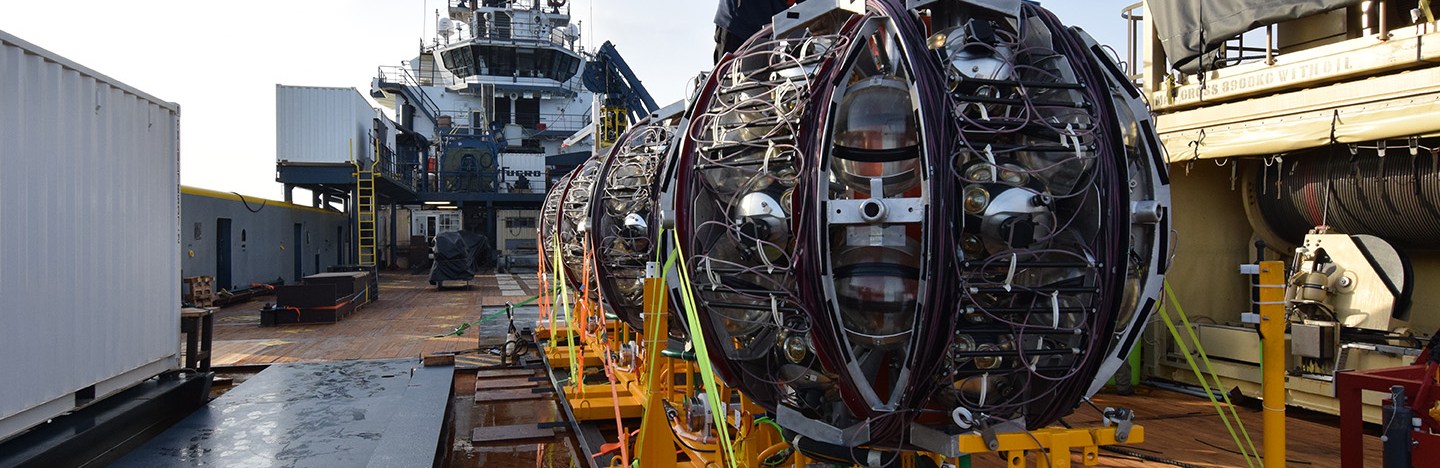 Workers on a ship prepare to launch a giant round piece of equipment off of a bright yellow ramp