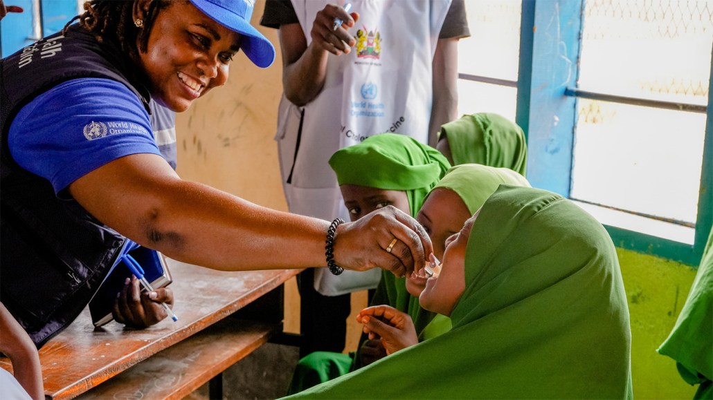 A WHO staff member wearing a blue hat and shirt gives a vaccine to girls in Kenya who are wearing green clothing. President Trump decided to withdraw the United States from the World Health Organization in January.
