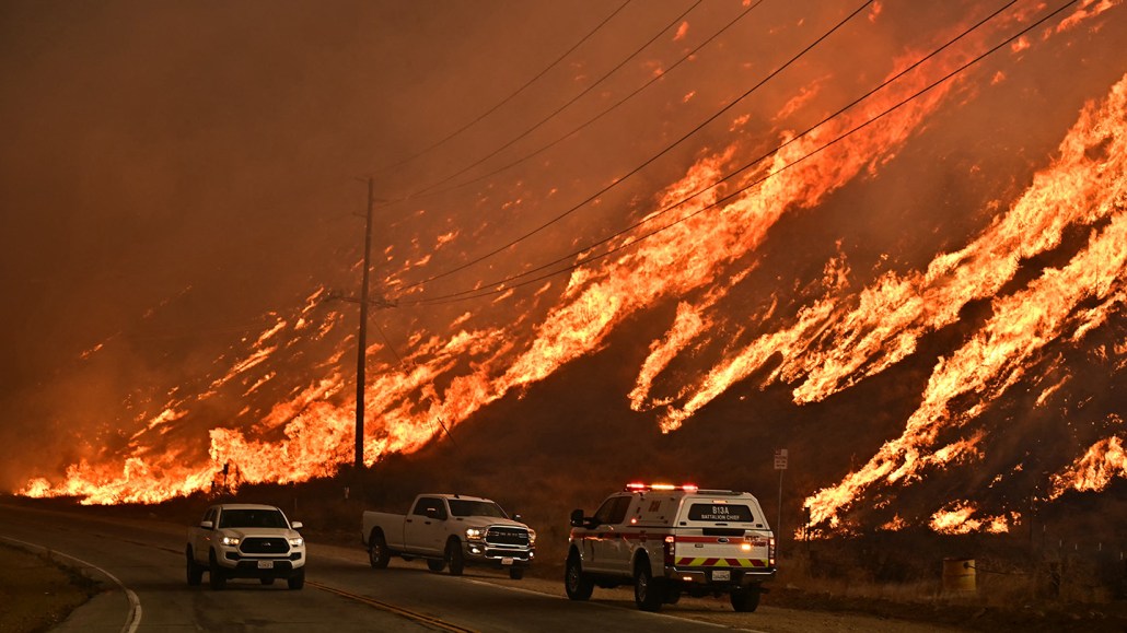 A wildfire burns on a hillside, as drivers in cars on a highway pass by in the foreground.