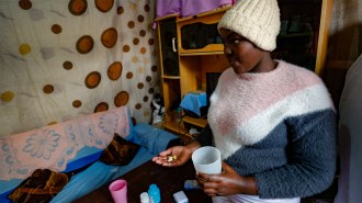 A Black girl with HIV is wearing a knit hat and a fuzzy pink, white and navy sweater and holding pills in her hand.