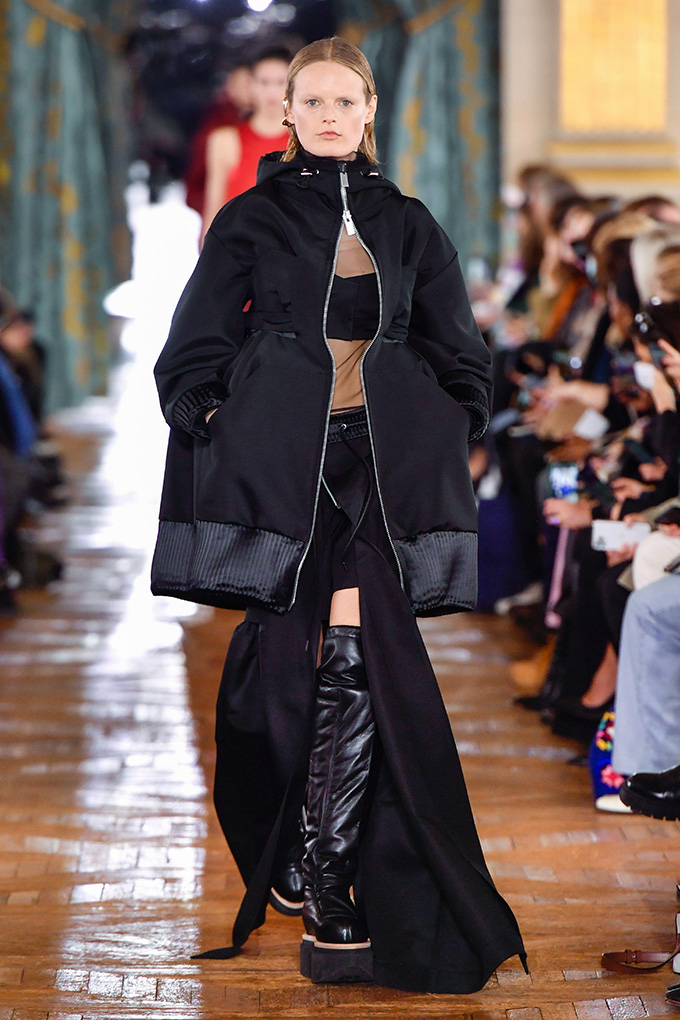 Model Hanne Gaby Odiele walks a runway with slicked back blonde hair and wearing an oversize black jacket, a tan top with a large black stripe across the chest, a black skirt with a thigh-high slit and chunky black thigh-high leather boots. Odiele is intersex.