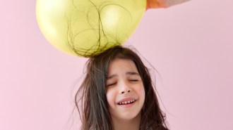 A girl closes her eyes as her long brown hair clings to a yellow balloon touching the top of her head.