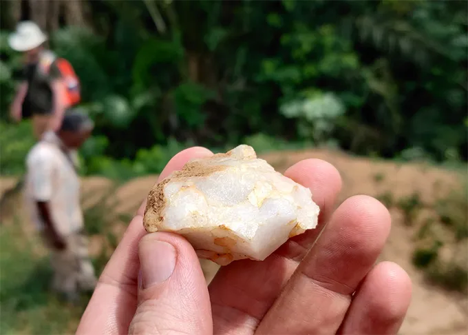 A photo of a hand holding a white stone in front of dense green foliage with two people out of focus in the background