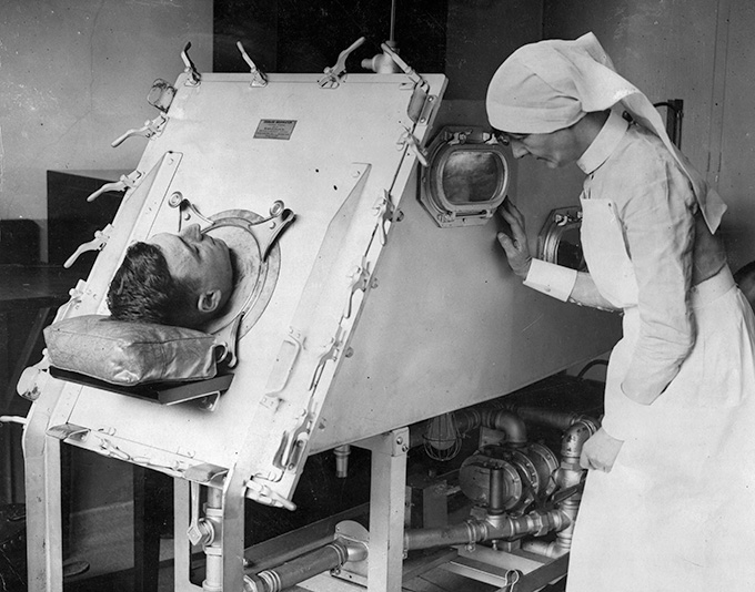 In this black-and-white image from 1938, a man's head sticks out of a large metal medical contraption called an iron lung, while a nurse peers through a window in the box.