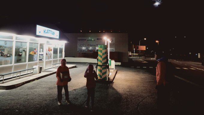 A photo of two people in the dark. They are carrying a box and standing in front of a gas station with a brightly-lit sign.