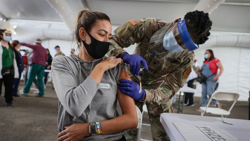 A U.S. Army soldier wearing a mask and face shield injects a vaccine into a woman's arm.