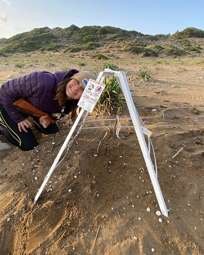 A woman kneels on the ground, leaning over a mound on a sandy landscape. The mound has a white tripod over it with some signage attached.