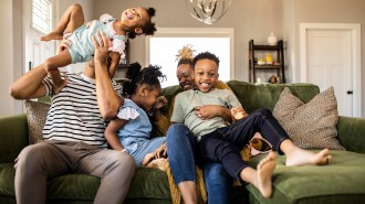 A mom and dad sit on a green couch holding their three kids playfully.