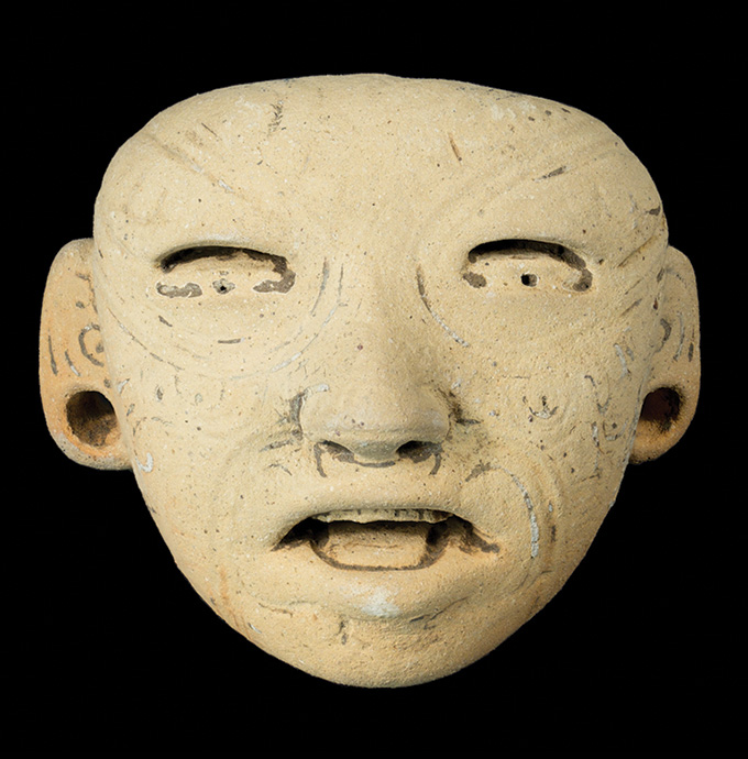 A close-up of an ancient clay face with a neutral expression, displayed against a black background. The face has hollowed-out eyes, an open mouth and circular markings or carvings on its surface. The ears are adorned with large, round perforations.
