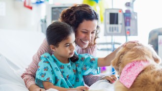 A girl, wearing patient attire, on a hospital bed pets a therapy dog. The girls' mother is sitting behind her smiling at the dog.