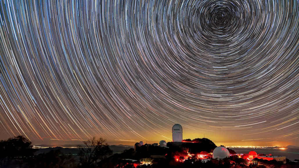 Stars swirl across the sky, creating a circular pattern in this time-lapse photo. In the foreground on a small rise of land is the Mayall Telescope at Kitt Peak National Observatory in Arizona, which is conducting the DESI dark energy survey.